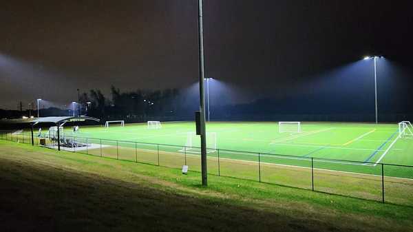 Lit sports field at night with soccer goals, fencing, and tall light poles illuminating green turf.